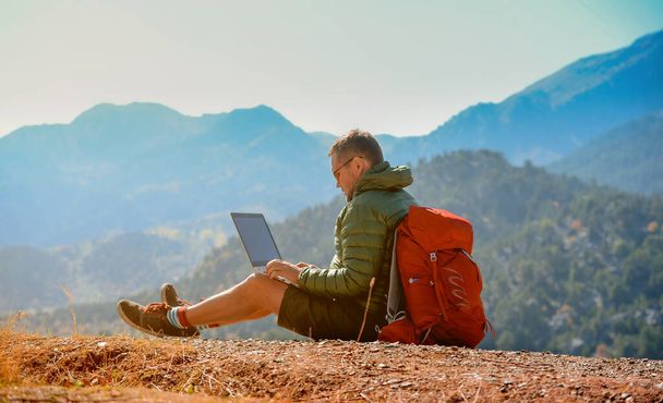 Человек-турист с ноутбуком, работающим на вершине гор. Удаленная работа с ноутбуком. Человек сидит на вершине - Фото, изображение