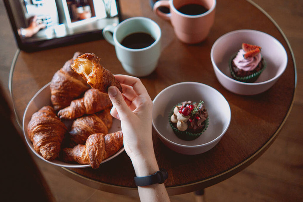 A woman picking croissant bread on white bowls with cupcake and coffee on a table - Foto, afbeelding