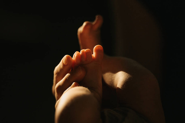A close-up photo of the gentle hand of the mother who is massaging the foot of her newborn baby. The legs of the infant in the sunlight in the evening. - Foto, Imagem