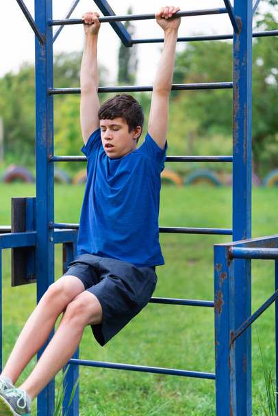 teenage boy exercising outdoors, sports ground in the yard, he hangs on the horizontal bar, raises his legs and pumps his abs, healthy lifestyle - Photo, Image