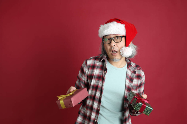 Старший азіатський чоловік носить червону сорочку з пластівцями, а капелюх Санта Клауса стоїть на червоному фоні.. - Фото, зображення