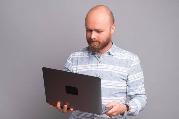 Serieuze man met baard in shirt met behulp van laptop over grijze achtergrond. - Foto, afbeelding