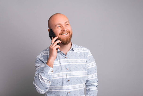 Smiling and joyful young man with beard talking on smartphone over grey background. - Fotoğraf, Görsel