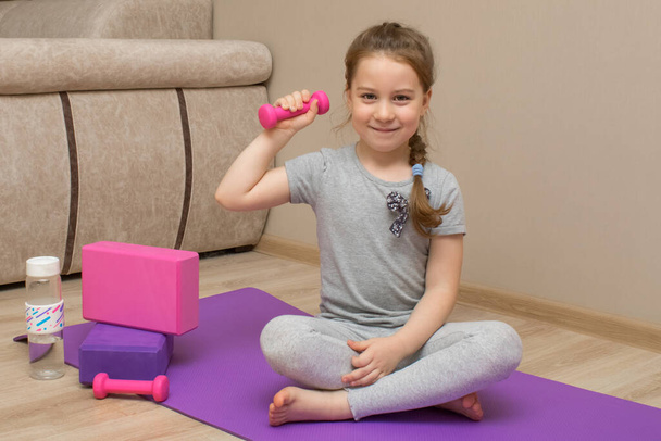 A little smiling girl sits on a fitness mat and holds a kettlebell in her right hand. She plays sports at home. Next to her are yoga blocks and a bottle of water. - Photo, image
