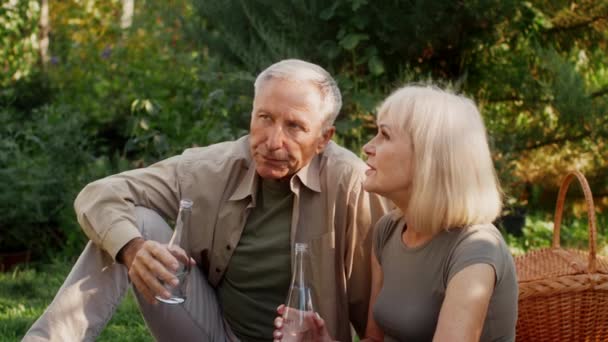 Gli sposi anziani che si rilassano all'aperto in giardino, bere acqua da bottiglie e chiacchierare, felice coppia matura che fa un picnic all'aperto, godendo di trascorrere del tempo sulla natura insieme, Filmati di rallentamento - Filmati, video