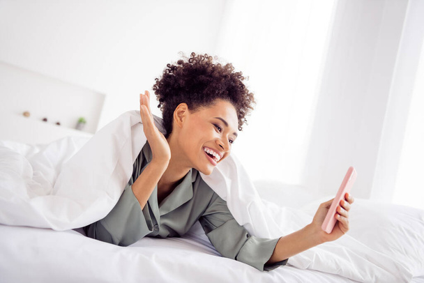 Retrato de menina de cabelos ondulados alegre atraente deitado na cama usando gadget chamando vídeo acenando Olá na casa casa luz dentro de casa - Foto, Imagem