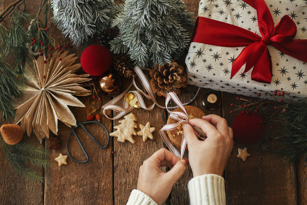 メリークリスマス！素朴な木製のテーブルの上にスタイリッシュなお祝いの装飾を背景にクリスマスオートミールクッキーにリボンを結ぶ手。健康的なクッキーとクリスマスの装飾と雰囲気のクリスマスイメージ - 写真・画像