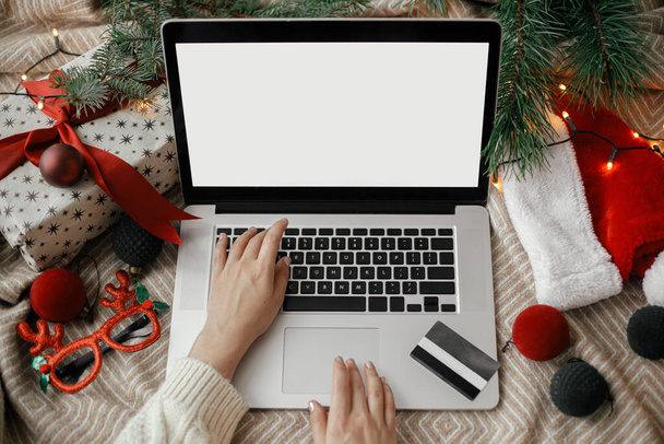 Manos en el ordenador portátil con pantalla en blanco en la cama acogedora con tarjeta de crédito, adornos de Navidad con estilo, presente, sombrero de santa. Venta de Navidad y compras en línea. Espacio para el texto. Vista arriba - Foto, imagen