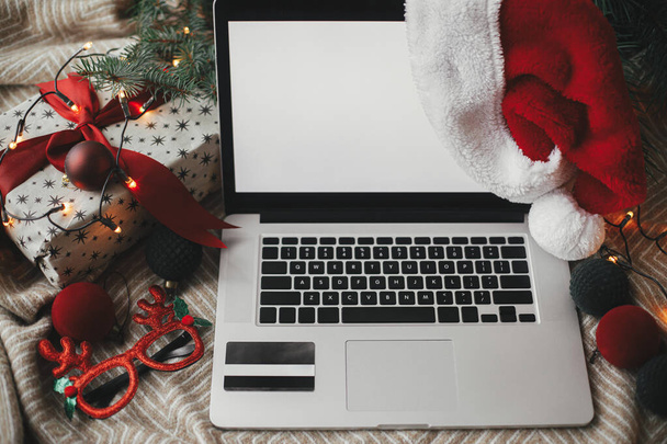 Boş ekranlı dizüstü bilgisayar ve rahat yatağında kredi kartı, şık Noel süsleri ve hediyeli Noel Baba şapkası. Noel satışları ve online alışveriş. Mesaj için yer var. Yukarıya bak - Fotoğraf, Görsel