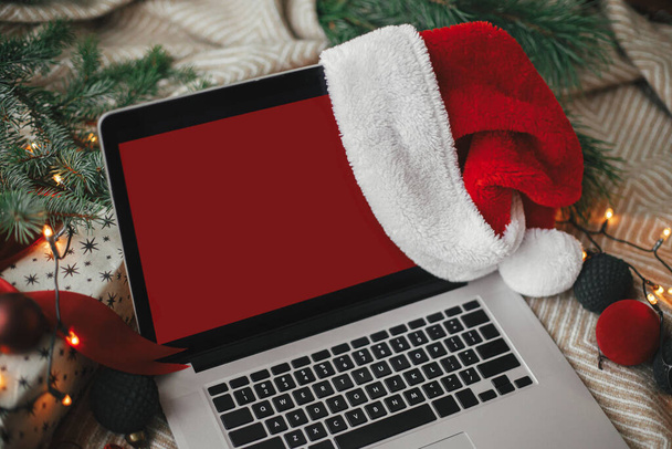 Laptop s červenou prázdnou obrazovkou v Santa klobouku na útulné posteli se stylovými vánočními ozdobami, současnými a slavnostními světly. Vánoční prodej a nakupování online. Prostor pro text. Pohled nahoru. Nezávislá - Fotografie, Obrázek