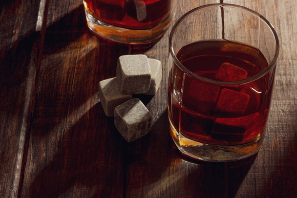 Transparent glass with whiskey on a dark wooden background with bright rays of light. Whiskey stones in the grocery - Фото, зображення