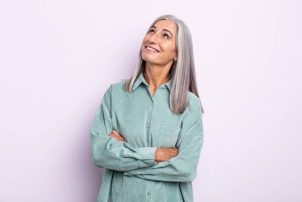 middle age gray hair woman feeling happy, proud and hopeful, wondering or thinking, looking up to copy space with crossed arms - 写真・画像