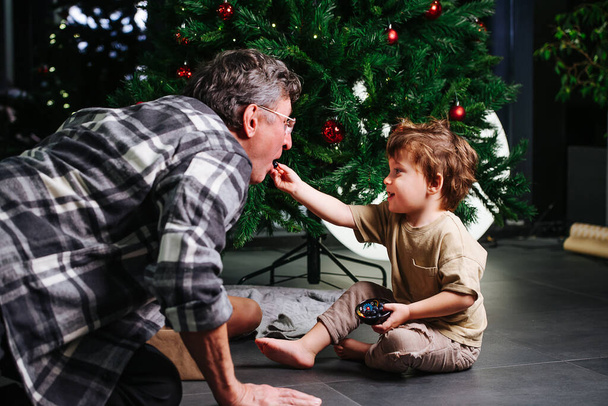Little boy feeding his grandpa standing on all four with some berries. In front of decorated christmas tree. - Foto, immagini