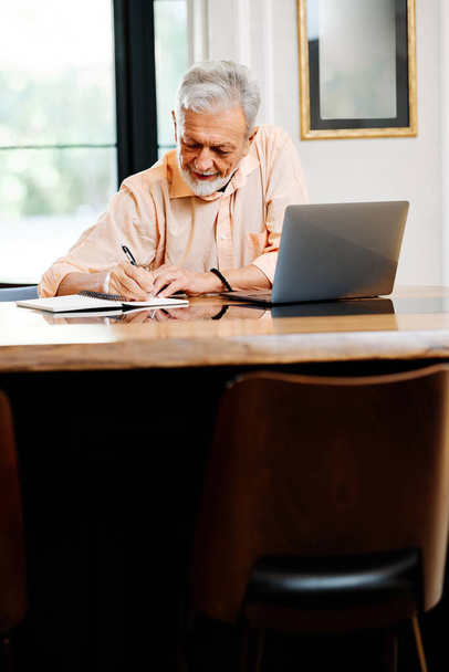 Un estudiante mayor feliz está sentado en casa y escribiendo notas en un cuaderno mientras sigue una conferencia en línea en el portátil. - Foto, Imagen