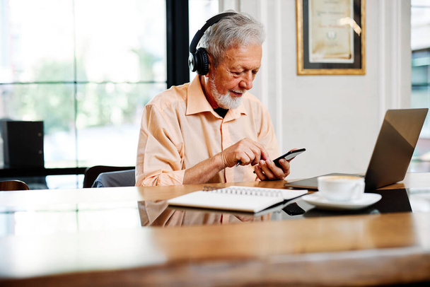 Oddaný seniorský student sedí v jídelně s nasazenými sluchátky a poslouchá online přednášku na notebooku. - Fotografie, Obrázek