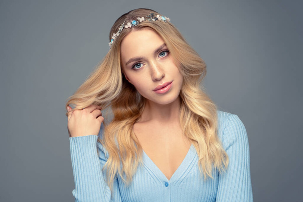 Portrait of a beautiful blonde with curly  long hair on a gray background with an ornament of small stones on her head and in a blue cardigan - Zdjęcie, obraz