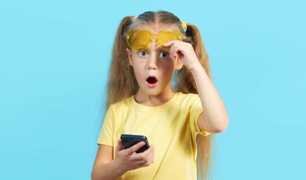 Awesome mobile app. A little surprised child girl in shape sunglasses pointing at cell phone with shocked face, online service, Parental control app. Studio shot blue background - Foto, Imagen