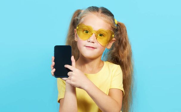 Awesome mobile app. A little surprised child girl in shape sunglasses pointing at cell phone with shocked face, online service, Parental control app. Studio shot blue background - Fotó, kép