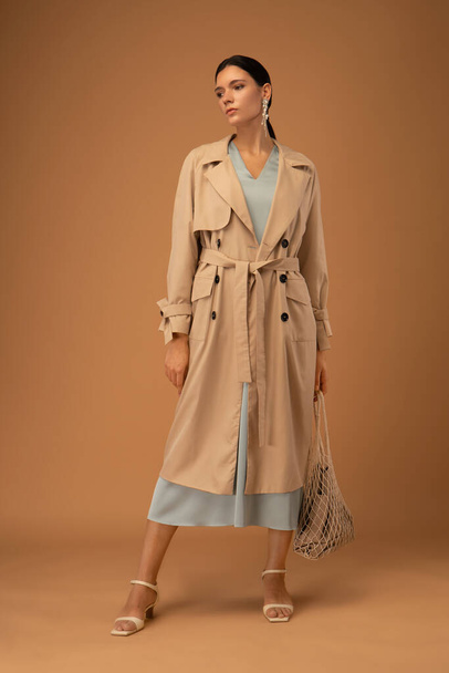 Elegant slim and tall brunette woman dressed in a blue dress and beige trench coat on a beige background - Fotó, kép