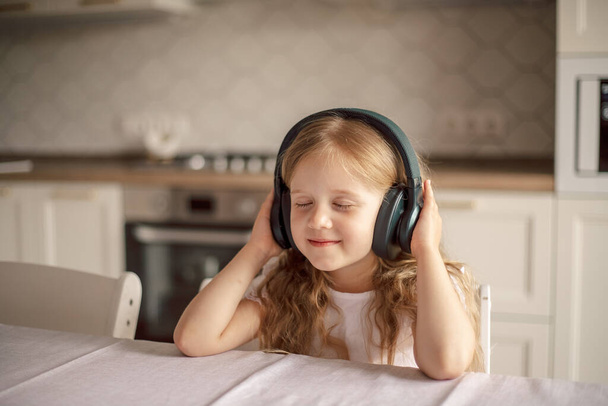Smiling cute little girl at home in headphones listens to music, audio. Child in the kitchen . kids and gadgets, blogger, streaming. technology concept. Music lessons, classical music - Photo, Image