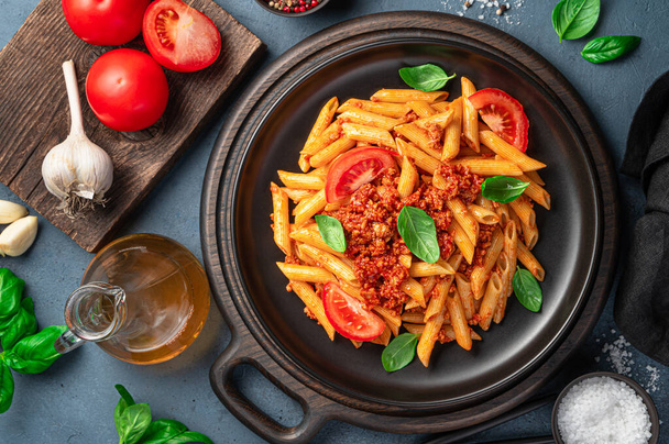 Penne pasta met bolognese saus in een zwarte plaat op een achtergrond van knoflook, tomaten en basilicum. Bovenaanzicht, close-up. - Foto, afbeelding