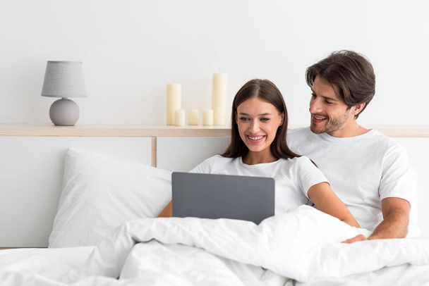 Smiling attractive young caucasian husband with stubble hugs wife sitting on bed, use laptop to surf in internet - Fotoğraf, Görsel
