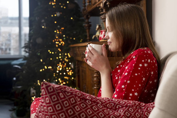Woman with tea cup in hands, sitting in chair at cozy home with fir tree on Christmas holidays - Foto, immagini