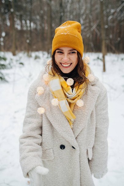 Smiling girl on Christmas Eve, walking in the winter forest, she put on a garland with lights on the top of the scarf.  - Foto, Imagen