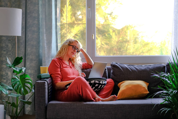 Ξανθιά γυναίκα με γυαλιά μιλάει στο τηλέφωνο κρατώντας τον φορητό υπολογιστή της κάθεται στον καναπέ στο παράθυρο στο σπίτι. - Φωτογραφία, εικόνα