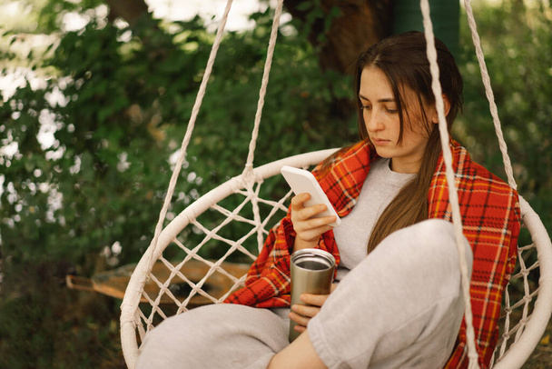 Meisje gewikkeld in geruite thee drinken en telefoon gebruiken in een hangstoel buiten. Mensen gebruiken technologie - Foto, afbeelding
