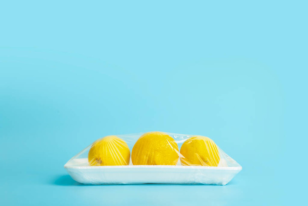 Fruits in plastic packaging from the supermarket are minimal. Lemons in cellophane and non-degradable plastic on a blue background. Biodegradable product packaging, environmental protection, nature - Foto, Imagen