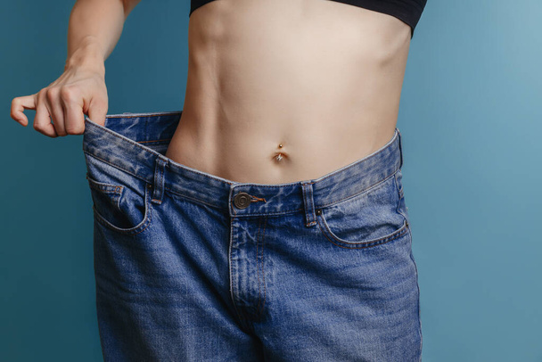 Attraktive, schlanke junge Frau in ihren Dreißigern, in weiten Jeans und weißem Hemd, demonstriert erfolgreiche Gewichtsabnahme. Das Konzept der Ernährung und Gewichtsabnahme.. Fettabsaugung. - Foto, Bild