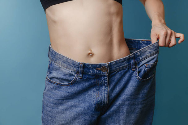 Привлекательная, стройная молодая женщина лет тридцати, одетая в широкие джинсы и белую рубашку, демонстрирует успешную потерю веса. Концепция диеты и потери веса... - Фото, изображение