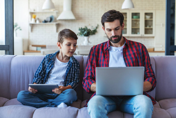 Feliz niño jugando en la tableta y su padre alegre trabajando en el ordenador portátil sentado en el sofá en casa. - Foto, Imagen