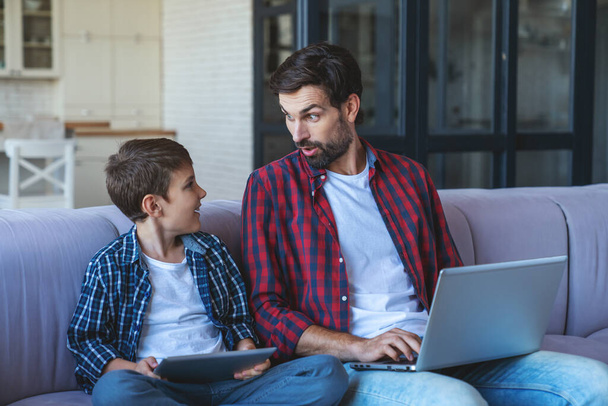 Szczęśliwy mały chłopiec bawiący się na tablecie i jego wesoły tata pracujący na laptopie siedząc na sofie w domu. - Zdjęcie, obraz