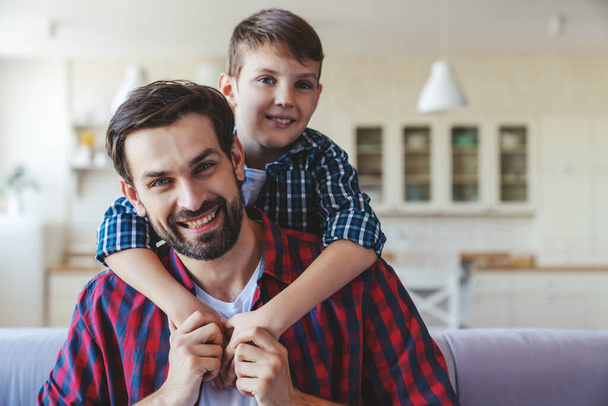 Маленький счастливый мальчик обнимает своего отца за шею, сидя дома на диване. - Фото, изображение