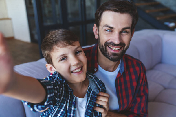 Happy little boy and his cheerful dad take a selfie sitting on the couch at home. - Foto, immagini