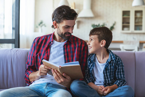 Happy little boy and his cheerful dad are reading a book sitting on the couch at home in the living room. - Φωτογραφία, εικόνα