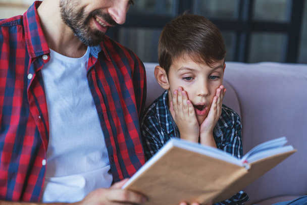 Happy little boy and his cheerful dad are reading a book sitting on the couch at home in the living room. - Фото, изображение