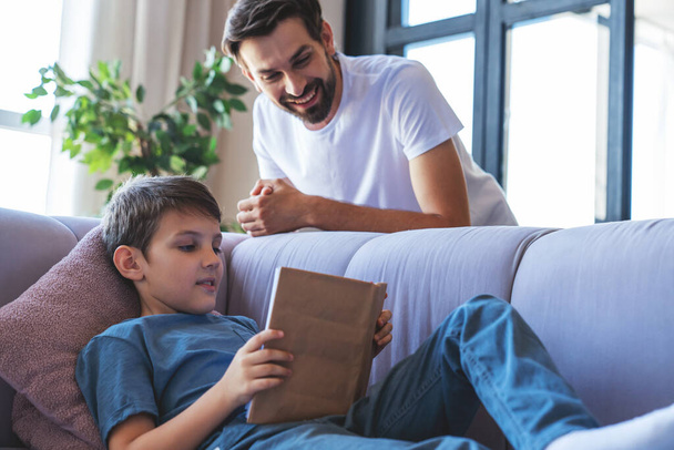 Щасливий маленький хлопчик і його веселий тато читають книгу, що сидить на дивані вдома у вітальні
. - Фото, зображення