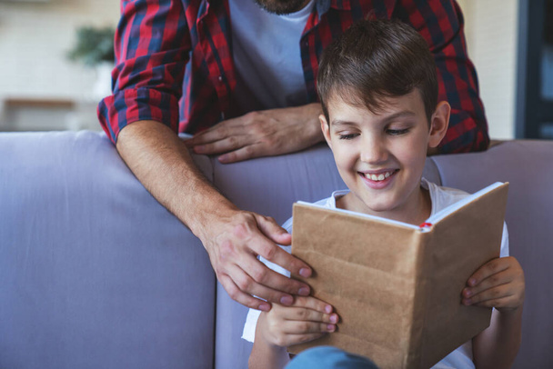 Happy little boy and his cheerful dad are reading a book sitting on the couch at home in the living room. - Foto, immagini
