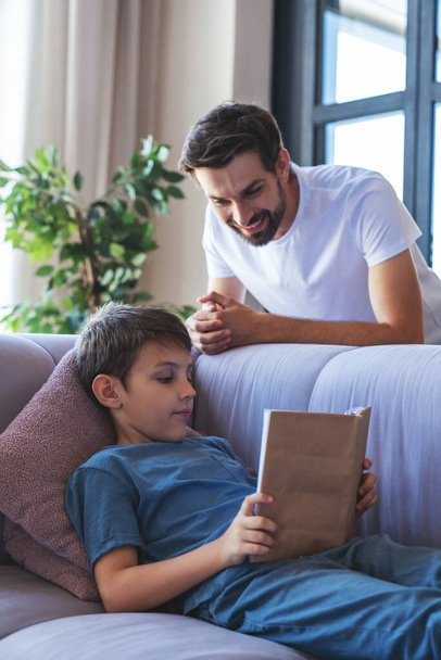 Feliz niño y su padre alegre están leyendo un libro sentado en el sofá en casa en la sala de estar. - Foto, imagen