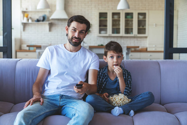 A happy little boy and his cheerful dad are sitting together on the couch in front of the TV, eating popcorn and cheerfully cheering for their favorite football team at home in the living room. - Фото, зображення