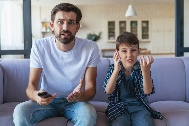 A happy little boy and his cheerful dad are sitting together on the couch in front of the TV, and cheerfully cheering for their favorite football team at home in the living room. - Фото, зображення