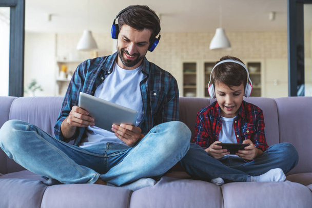 Happy Little Boy ja hänen iloinen isä kuuntelevat musiikkia kuulokkeilla, käyttäen vempaimia istuu sohvalla kotona olohuoneessa. - Valokuva, kuva