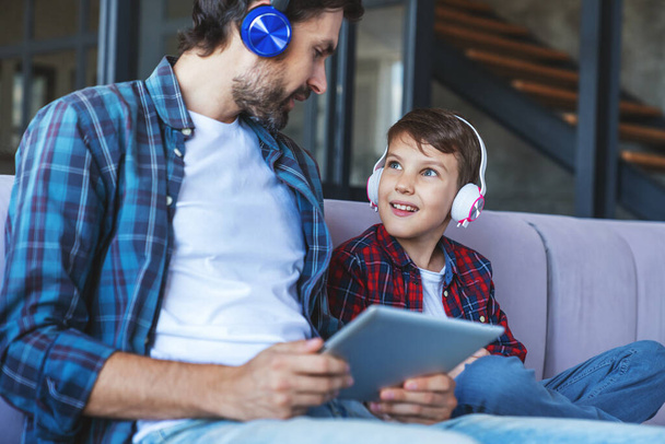 Szczęśliwy chłopczyk i jego wesoły tata słuchają muzyki na słuchawkach, używając gadżetów siedzących na kanapie w domu w salonie. - Zdjęcie, obraz