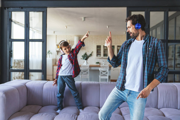 Ο χαρούμενος μπαμπάς και ο μικρός γιος του ακούνε μουσική με ακουστικά και χορεύουν στο σπίτι στο σαλόνι.. - Φωτογραφία, εικόνα