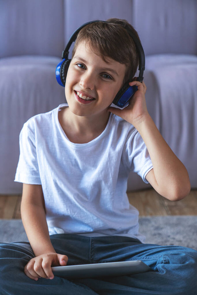 Портрет маленького хлопчика, який слухає музику на навушниках і використовує електронний планшет, що сидить на підлозі вдома у вітальні
. - Фото, зображення