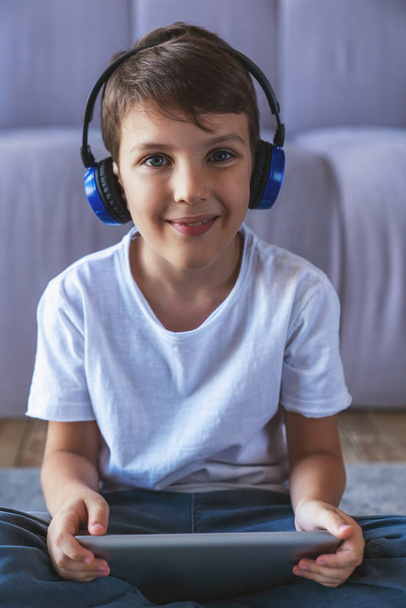 Portret van een jongetje dat naar muziek luistert op een koptelefoon en een elektronische tablet gebruikt die thuis in de woonkamer op de vloer zit. - Foto, afbeelding