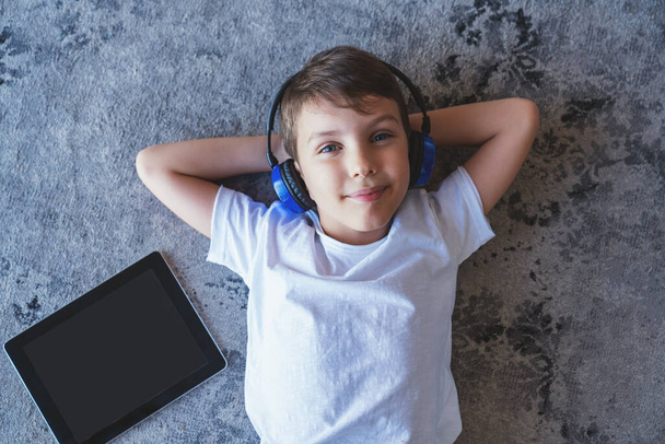Portrait of a little boy listening to music on headphones and using an electronic tablet lying on the floor at home in the living room. - Photo, Image